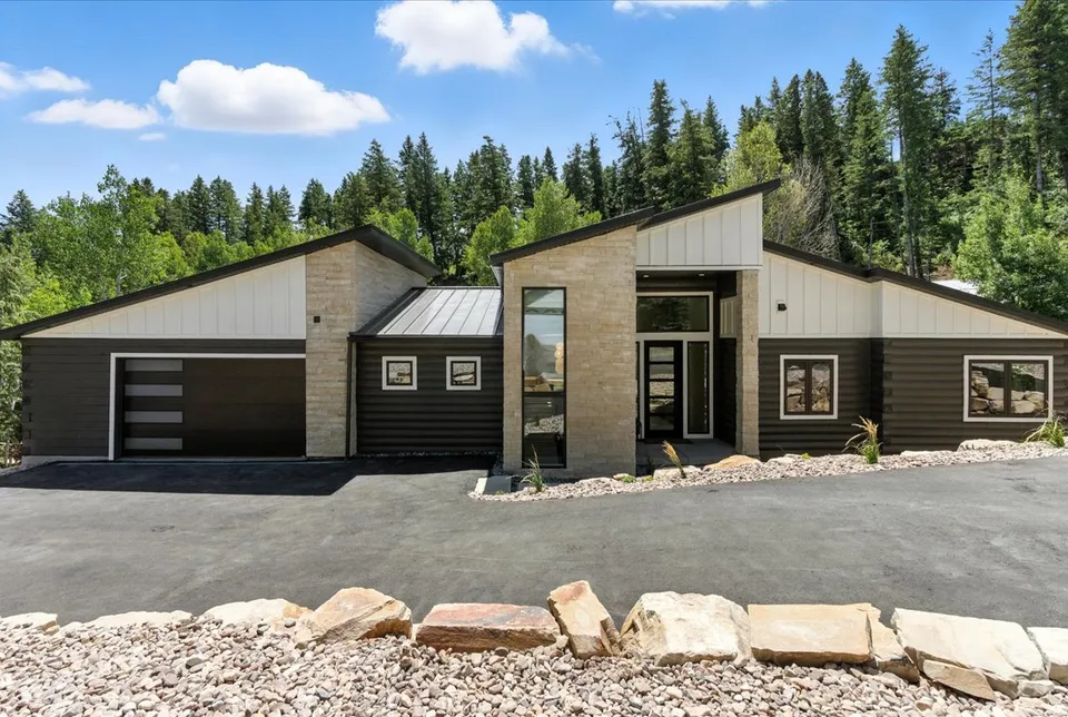 A modern mountain home with stone and dark wood siding and a glass front entry, set against a backdrop of tall evergreen trees.