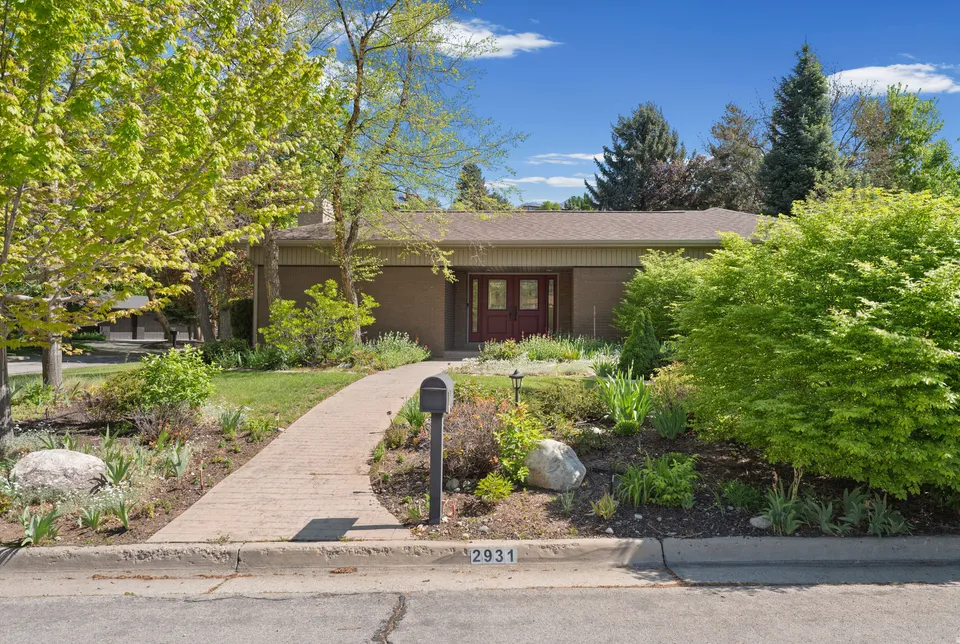 A single-story brick home with burgundy double doors and a brick front walkway, framed by mature trees and established garden beds.