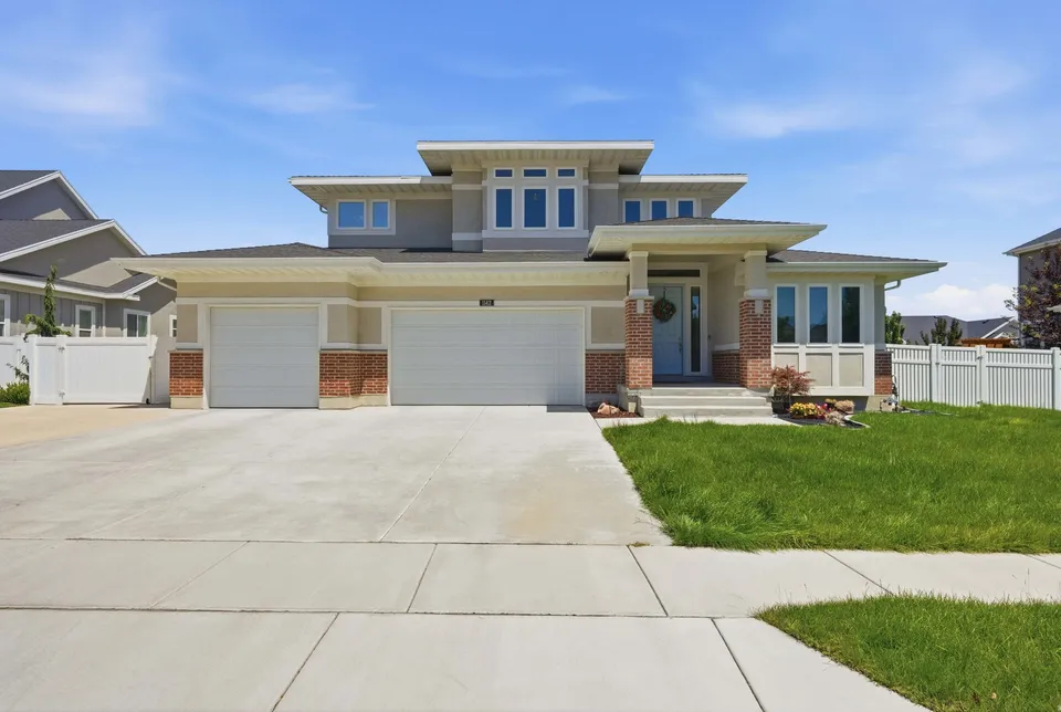A two-story stucco Prairie-style home with red brick accents, a three-car garage, and a manicured green lawn under a clear blue sky.